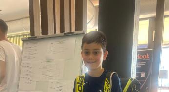 Ragazzo sorridente con zaino da tennis, accanto a un tabellone di torneo.