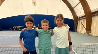 Tre bambini sorridenti abbracciati su un campo da tennis, pieni di energia e felicità.