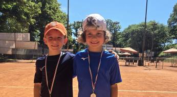 Due bambini sorridenti con medaglie su un campo da tennis soleggiato.